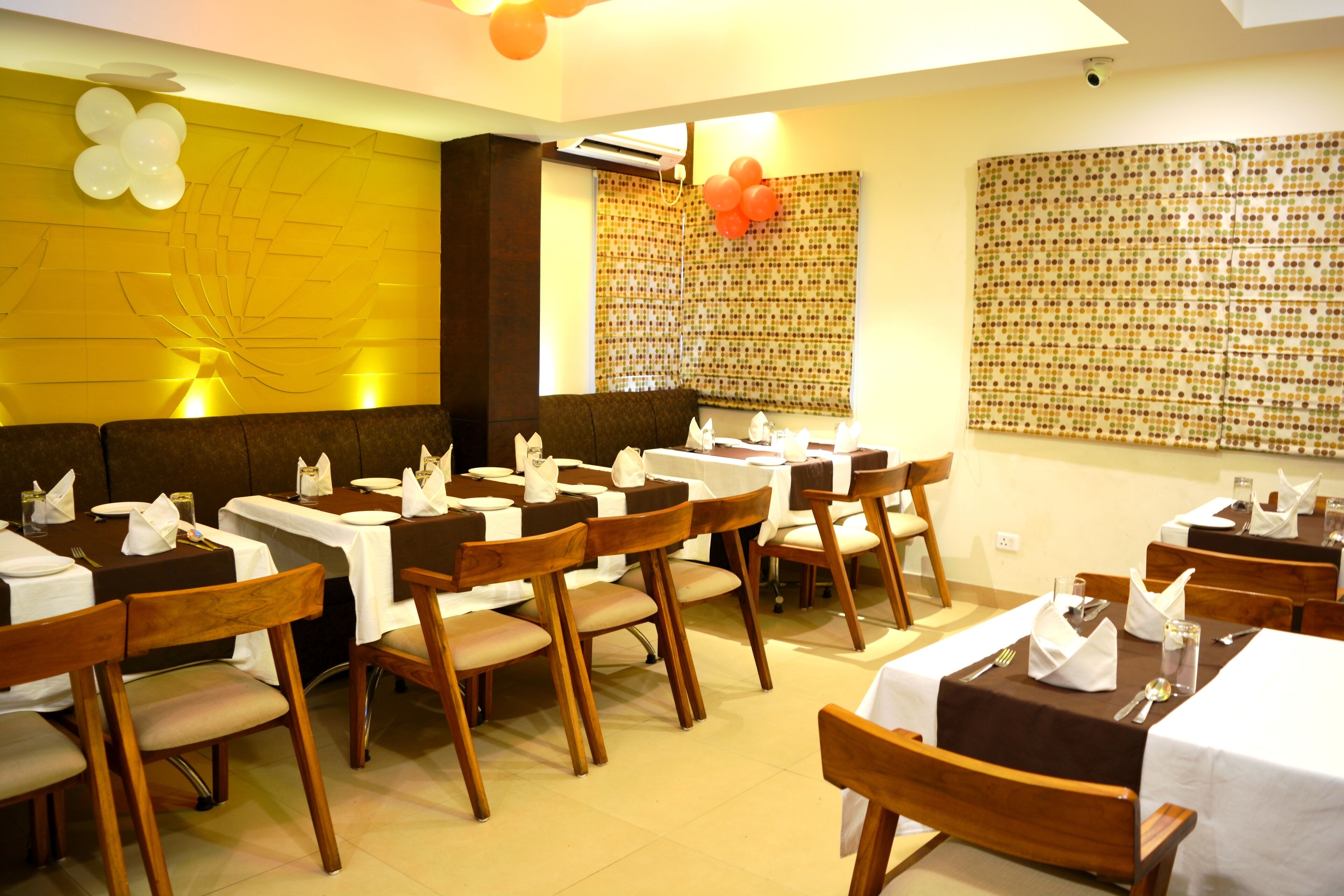 Un restaurant doté de nombreuses tables recouvertes de nappes blanches et entourées de chaises en bois, avec un mur d’accent jaune et un papier peint à motifs.