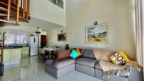 A spacious living area with a grey sectional sofa, a dining table with chairs, a kitchen with a refrigerator, and a staircase.