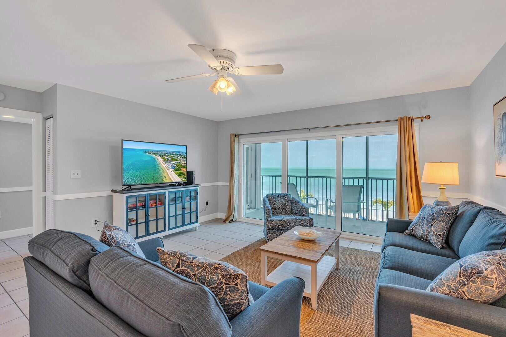 A living room with a television, sofas, a coffee table, and a balcony with a view of the ocean.