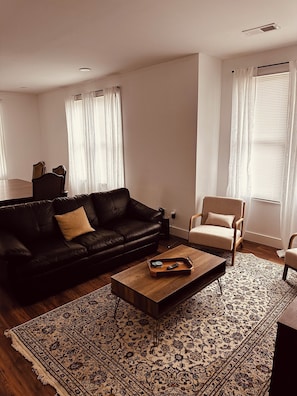 Een moderne woonkamer met een zwarte leren bank, een houten salontafel, een beige fauteuil en een kamerkleed met een patroon.