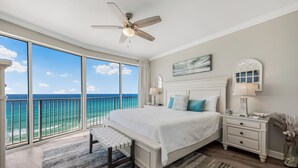 A bedroom with a large bed, a nightstand, a bench, a ceiling fan, and a view of the ocean.