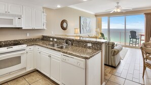 Una cocina con armarios blancos, microondas y lavavajillas. Hay una zona de comedor con un sofá a rayas y vistas al océano.