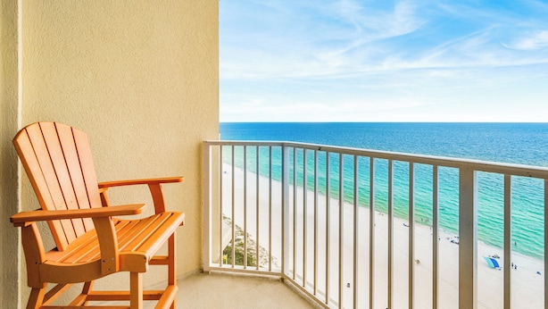 A wooden chair on a balcony overlooking a beach with clear blue water and a white sandy shore. - Upscale but Within Reach on the Beach! (Panama City Beach)