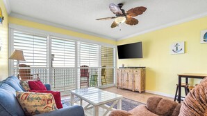 A living room with a sofa, armchair, and a coffee table. There is a ceiling fan, a TV mounted on the wall, and a window with blinds. - Upscale but Within Reach on the Beach! (Panama City Beach)