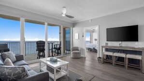 A spacious living area with a balcony overlooking the ocean, a flat-screen TV, and a dining area with stools.