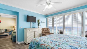 Un dormitorio con una cama grande, ventilador de techo, televisor, ventana con persianas y vista al océano.