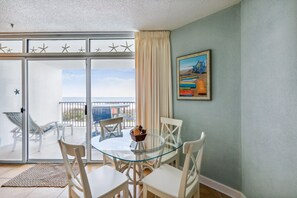 Quarto com mesa e cadeiras de vidro, um quadro na parede e vista para o mar através de uma porta de correr.