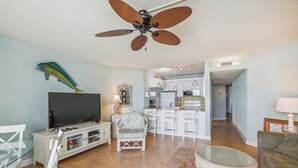 Un salon avec un téléviseur, des meubles blancs, un ventilateur de plafond et une cuisine à l’arrière-plan.