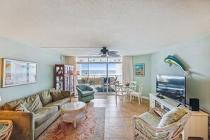 A living room with a sofa, a dining table, chairs, a TV, and a view of the ocean through large windows.