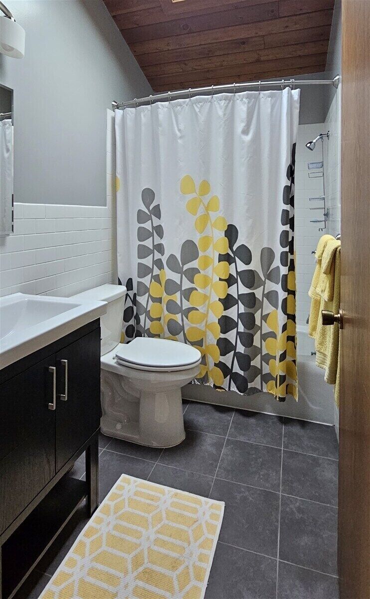 Un baño con inodoro blanco, cortina de ducha con estampado de hojas, techo de madera y toalla amarilla en un perchero.