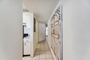 A narrow hallway with a kitchen area on the left, a white door with intricate designs on the right, and a tiled floor.