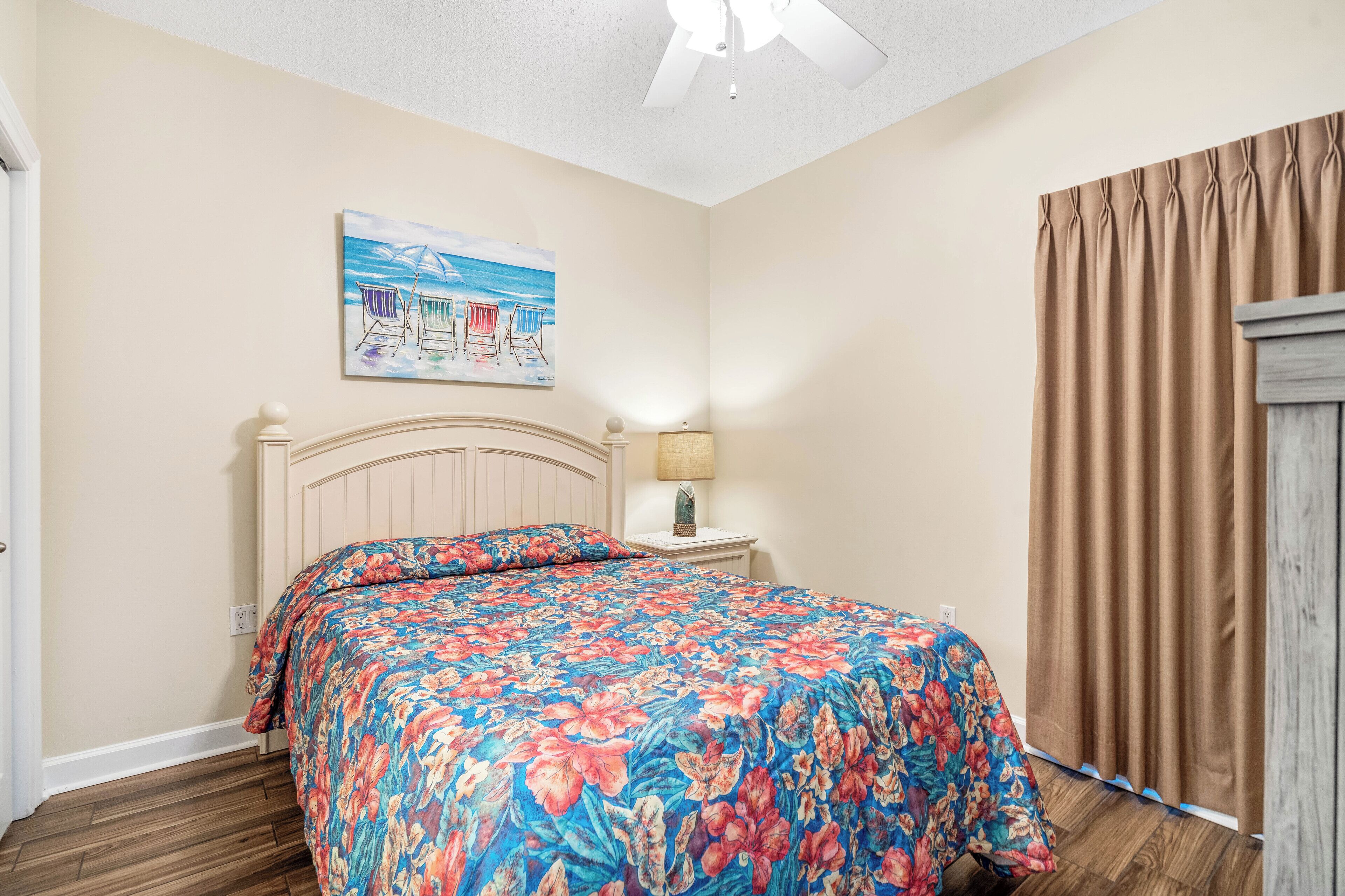 Une chambre à coucher avec une couvre-lit à motifs floraux, un tableau représentant des chaises de plage, un ventilateur de plafond et une table de chevet en bois avec une lampe.