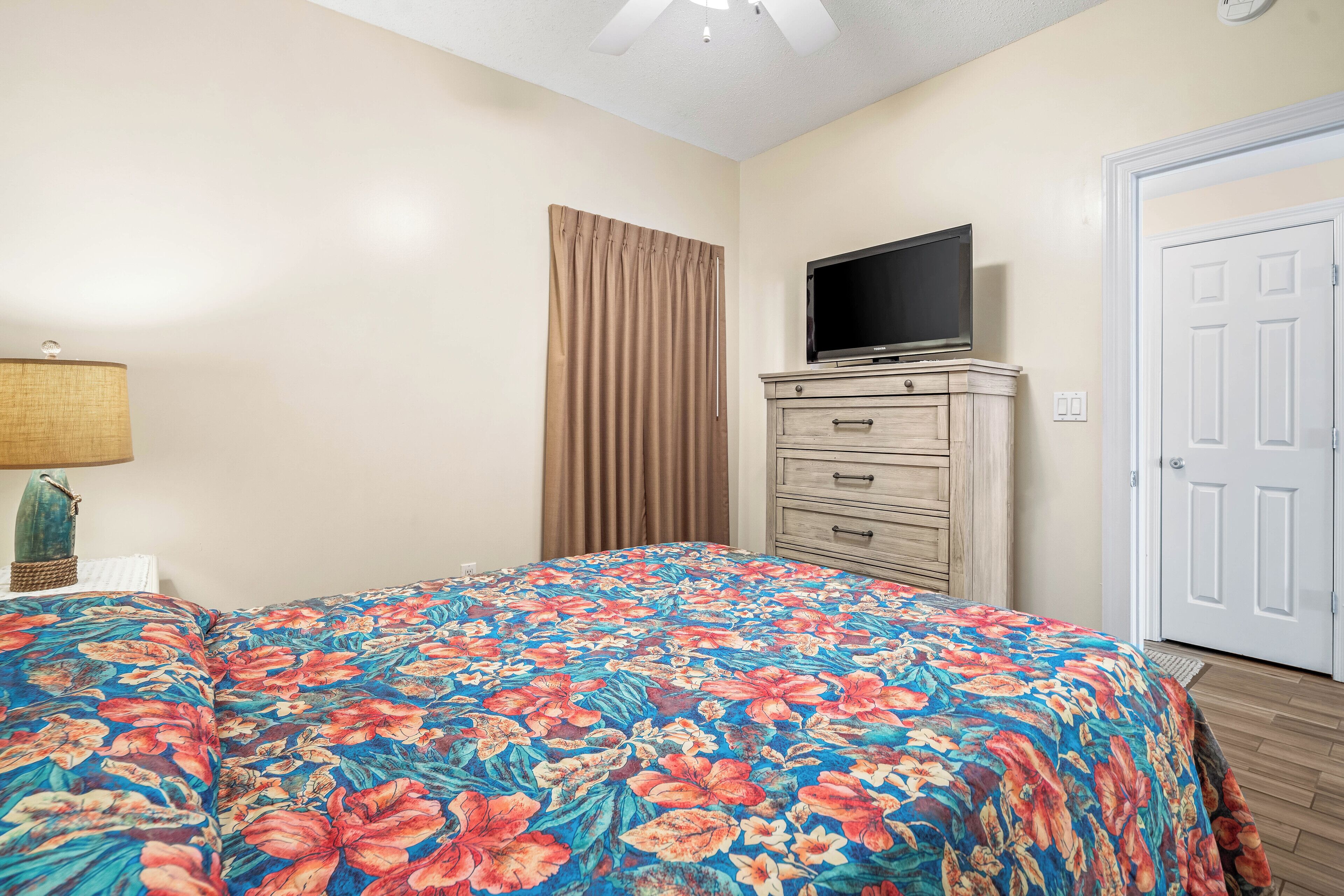 Un dormitorio con colcha estampada, un televisor sobre una cómoda, una puerta y una lámpara en una mesita de noche.