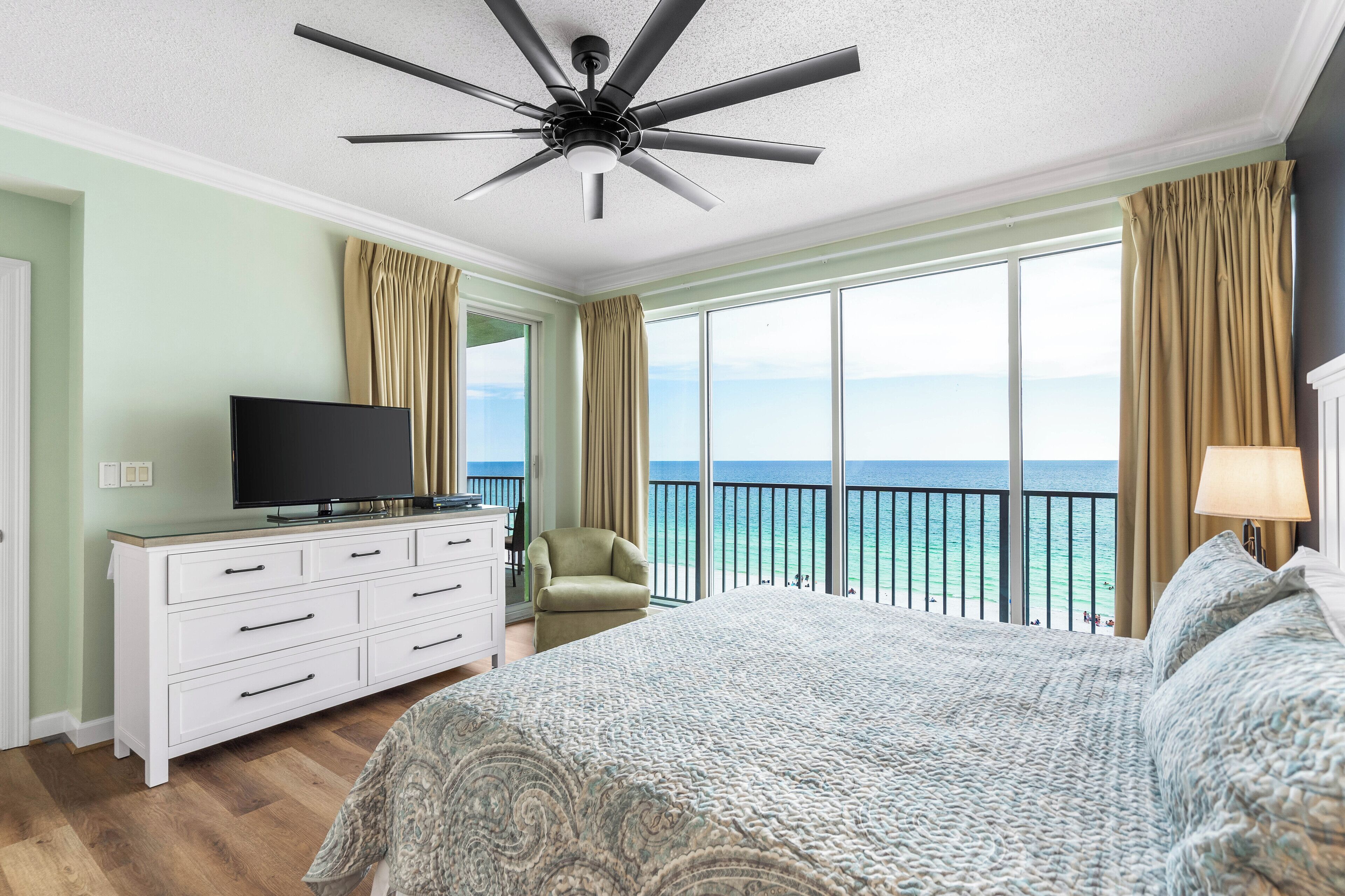 Una camera d'albergo con un letto grande, una TV su una cassettiera bianca, un ventilatore a soffitto e un balcone con vista sull'oceano.