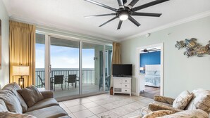 A spacious living area with a ceiling fan, a sofa, a patterned armchair, a television, and a sliding glass door leading to a balcony with a view. - Deluxe Beachfront Condo (Panama City Beach)