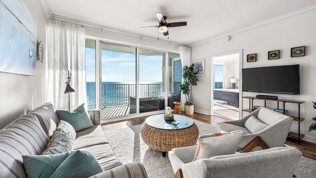 Un salon comprenant un canapé, des fauteuils, une table basse et un téléviseur. Il y a un balcon avec vue sur l’océan.