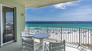 A balcony with white wicker furniture, a round table, and a view of a beach with umbrellas and people.