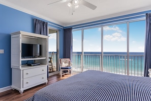 A bedroom with a large bed, a TV on a dresser, a chair, and a balcony with a view of the ocean.