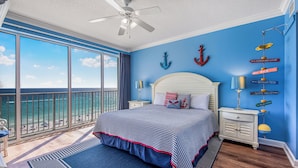 A bedroom with a large bed, a ceiling fan, a nightstand, a dresser, and a view of the ocean. - Deluxe Beachfront Condo (Panama City Beach)