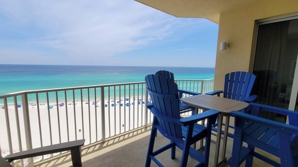 Ein Balkon mit zwei blauen Adirondack-Stühlen und einem kleinen Tisch, mit Blick auf einen Strand mit Sonnenschirmen und Menschen.