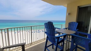 Varanda com duas cadeiras Adirondack azuis e uma mesa, com vista para uma praia com guarda-sóis e pessoas.