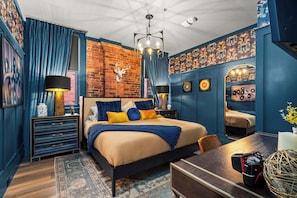 A bedroom with a brick wall, a bed with blue and yellow bedding, a dresser, a desk with a lamp, and a mirror.