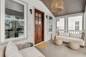 Een overdekte veranda met een witte gevel, een houten deur en een beige bank met kussens.