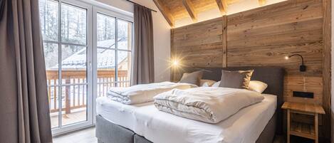 A bedroom with a large bed, wooden ceiling beams, a sliding glass door to a balcony, and a nightstand with a lamp.