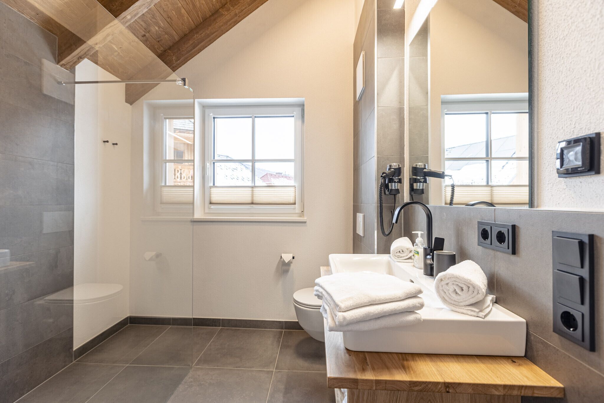 Een moderne badkamer met douche, toilet en wastafel. Er liggen handdoeken op de wastafel en hangt er een grote spiegel boven.