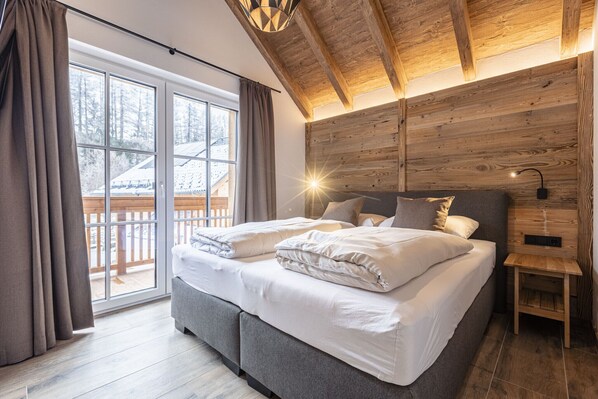 Une chambre avec un grand lit, des poutres apparentes en bois au plafond, une table de chevet et une vue sur une terrasse enneigée.