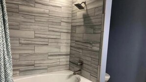 A bathroom with a white bathtub, a shower, and a toilet. The walls are covered with a grey marble-like tile pattern.