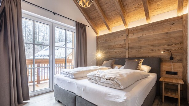 Une chambre avec un grand lit, des poutres apparentes en bois au plafond, une table de chevet et une vue sur une terrasse enneigée.