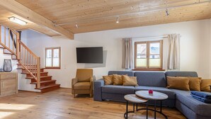 A modern living room with a wooden staircase, a grey sofa set, a small coffee table, and a flat-screen TV mounted on the wall.