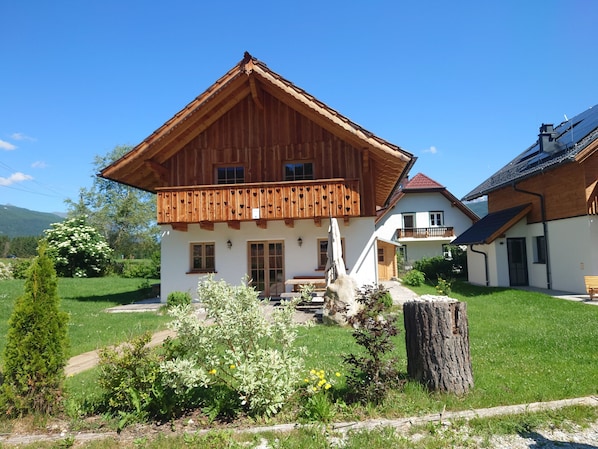 A two-story wooden house with a balcony, surrounded by a well-maintained garden.