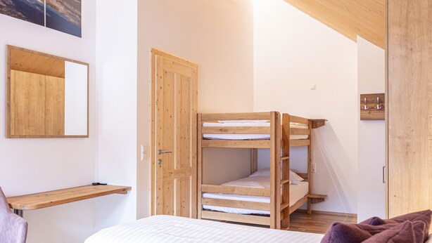Een stapelbed met houten frame, een wit matras en een hoofdbord. Links op de muur bevindt zich een houten plank, en rechts een deur met een houten frame.
