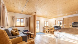 Ein geräumiger Wohnbereich mit einer Couch, einem Couchtisch und einem Esstisch. Im Hintergrund ist eine Küche mit Holzschränken und einer Arbeitsplatte zu sehen.