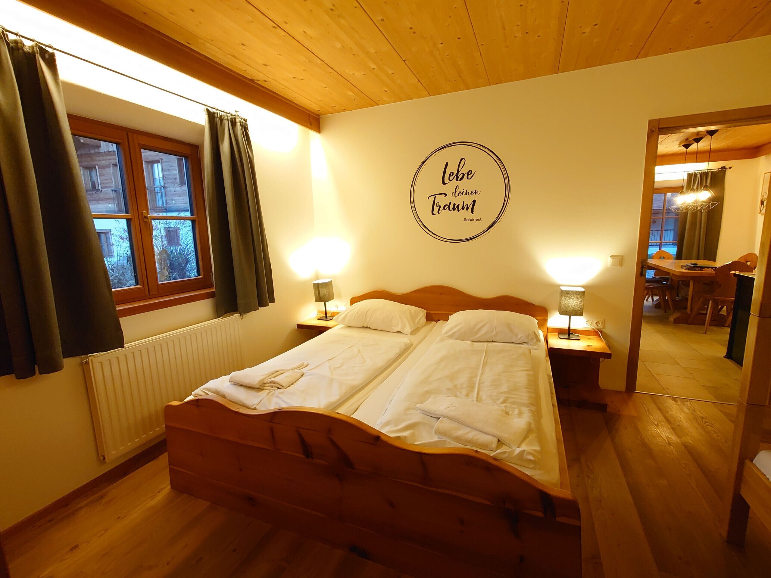 Um quarto aconchegante com uma cama de casal, duas luminárias de cabeceira, um encosto de madeira, uma janela com cortinas e uma placa na parede.