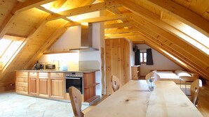 A spacious attic room with a wooden dining table, chairs, a kitchen area with wooden cabinets, and a large window providing natural light.