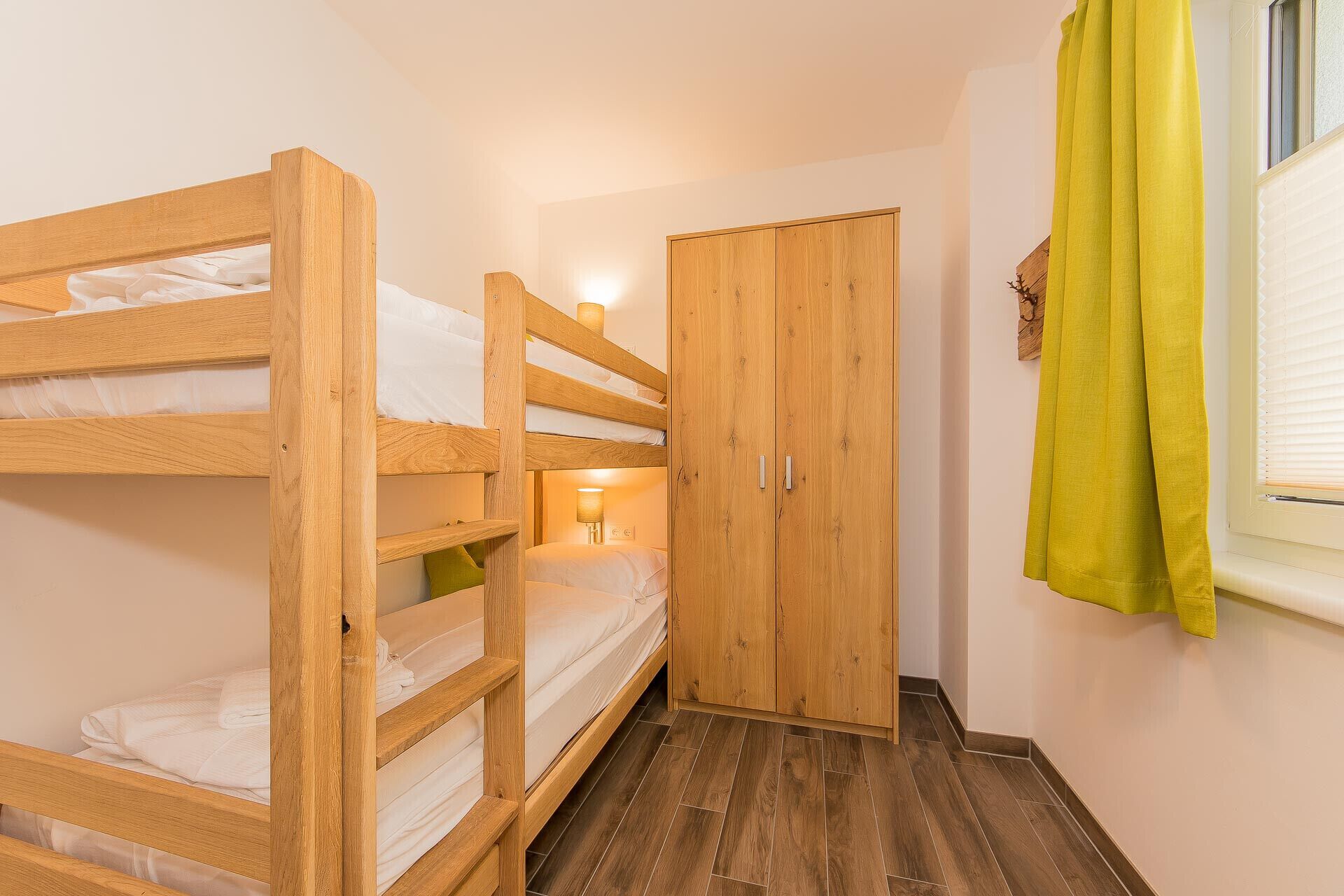 A bunk bed room with a wooden wardrobe and a window with curtains.
