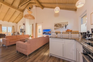 Amplia sala de estar con un sofá grande, una cocina con electrodomésticos modernos y un comedor con mesa y sillas de madera.