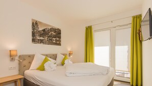 Una habitación de hotel con cama, mesita de noche, lámpara de pared, un cuadro y una puerta corrediza con cortinas.