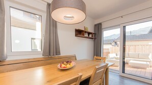 Ein Essbereich mit Holztisch, Stühlen, einer Obstschale, einer Hängelampe, einem Fenster mit Vorhängen und Blick auf einen Garten mit Holzzaun und Gartenmöbeln.