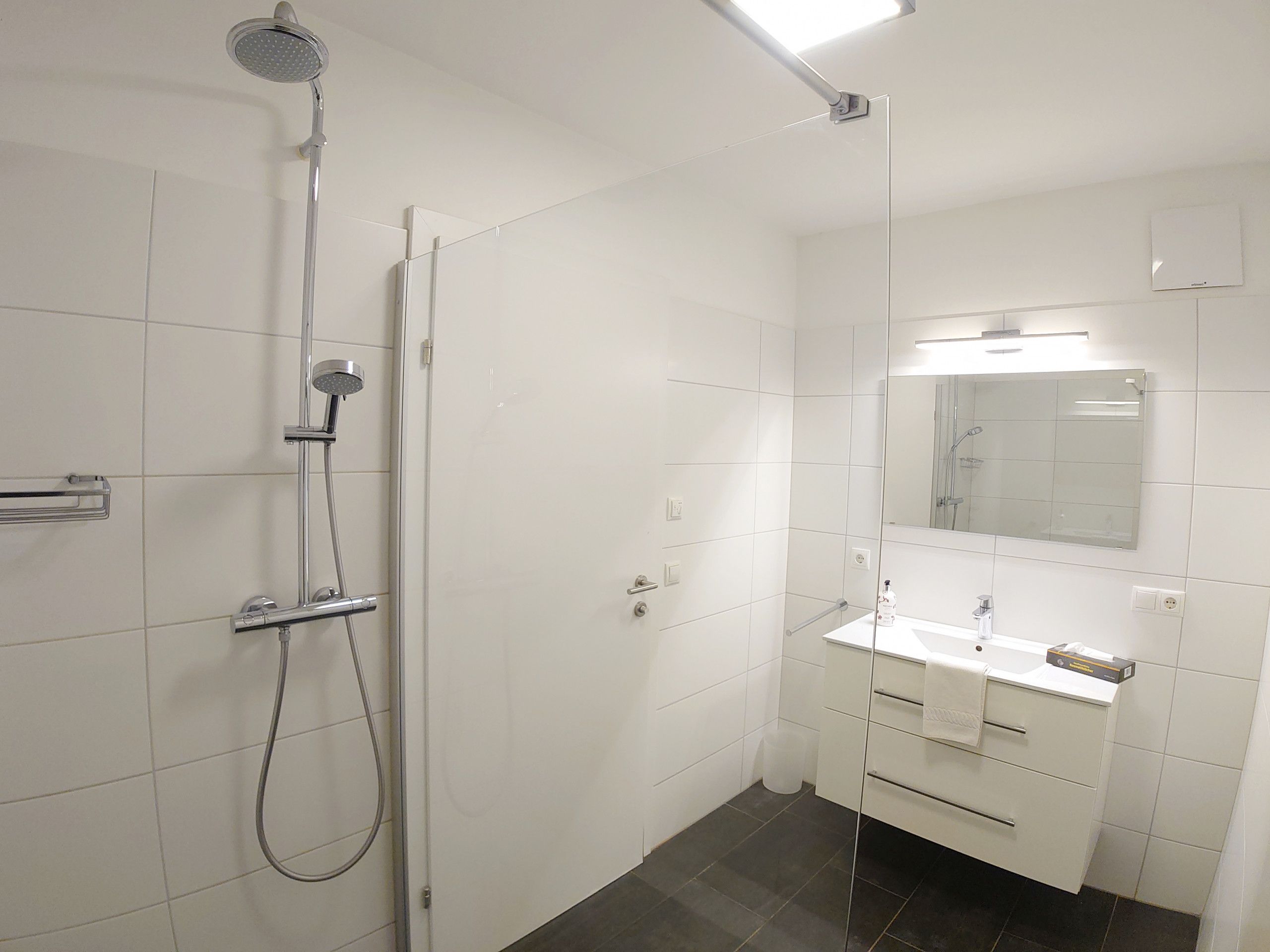 A modern bathroom with white tiles, a glass shower enclosure, a white sink with a mirror above it, and a dark floor.