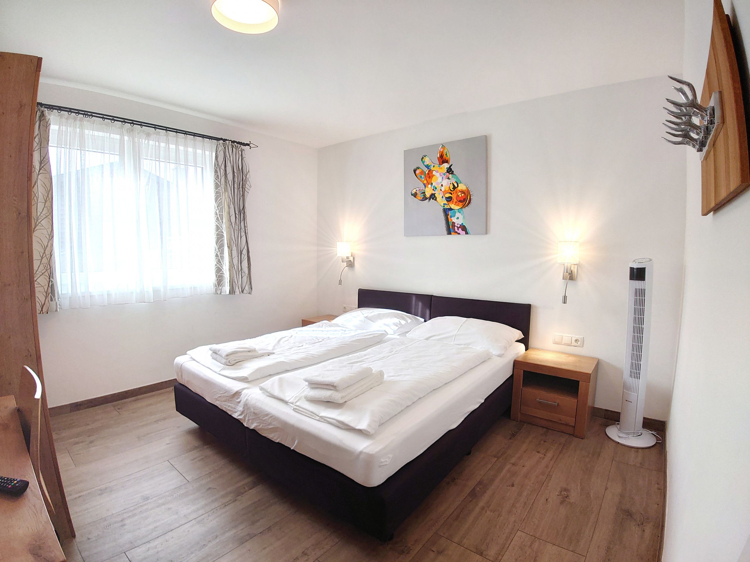 A neatly made double bed with white linens, a bedside table with a lamp, a wall-mounted artwork, and a standing heater.