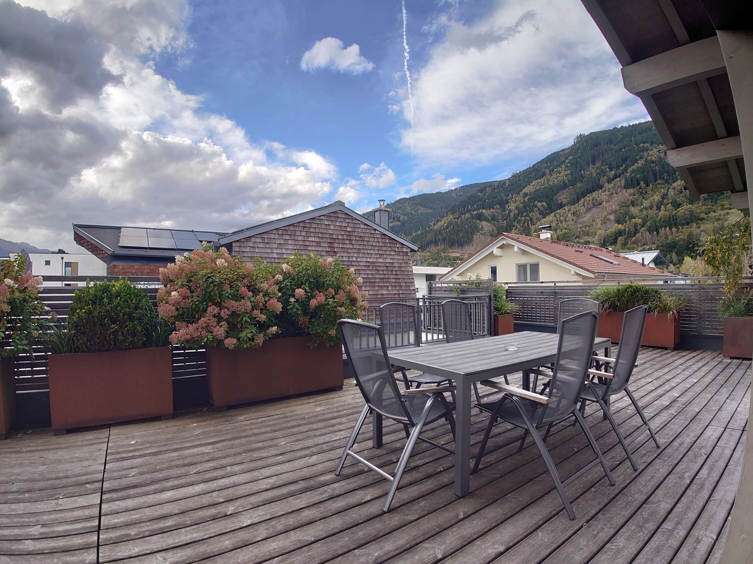 Eine Holzterrasse mit Tisch und Stühlen, umgeben von Pflanzkästen mit rosa Blüten, mit Blick auf eine gebirgige Landschaft.