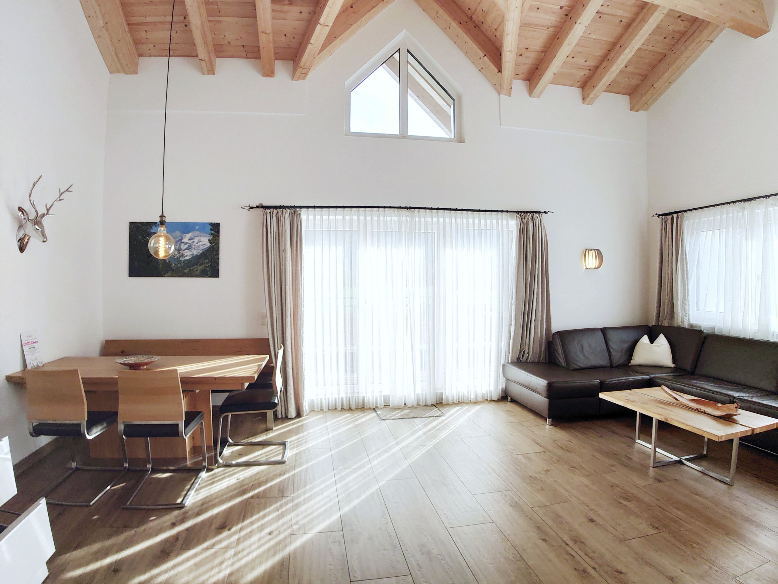 Een ruime woonkamer met houten plafond, een eettafel met stoelen, een bank en een groot raam met gordijnen.