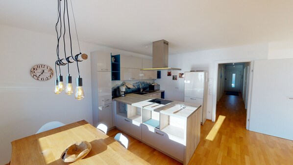 Une cuisine moderne avec des armoires blanches, un îlot central et une cuisinière intégrée. Il y a une horloge au mur et un luminaire suspendu au-dessus de l’îlot.