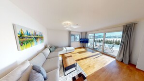A modern living room with a sofa, a coffee table, and a flat-screen TV. There is a large window with a view of a snowy landscape. - Kitzview Idyll Apartment (Zell am See)