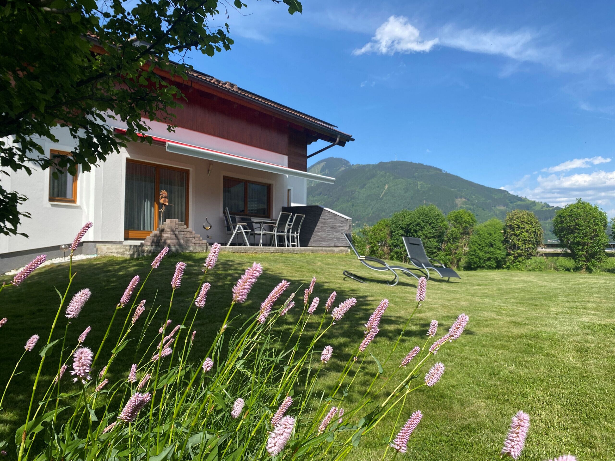 A modern house with a red roof, a patio with outdoor furniture, and a grassy lawn with pink flowers.
