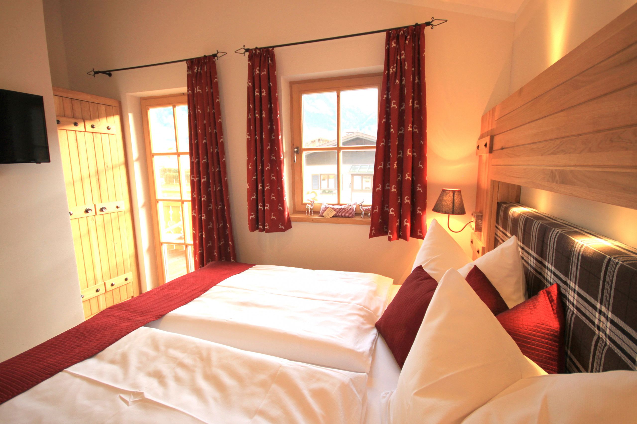 A hotel room with a bed, two windows, a wooden headboard, and a television on the wall.
